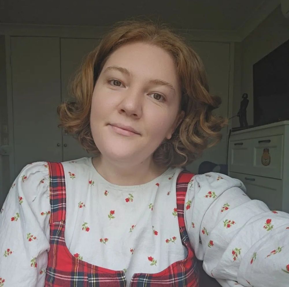 A selfie of Ella, who wears a white long sleeve shirt under a red tarten top. She has bob-length curly red hair, and is smiling at the camera.